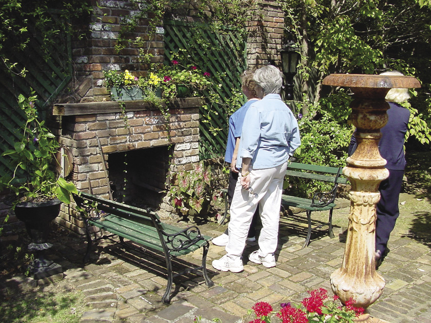 Three people stand on a brick patio looking at an outdoor brick fireplace. Two green benches face each other in front of the fireplace.